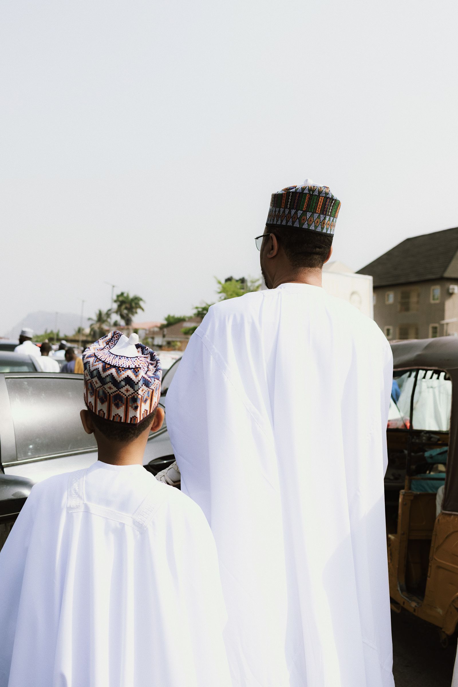 A father and son in white kaftans and embroidered caps, seen from behind.